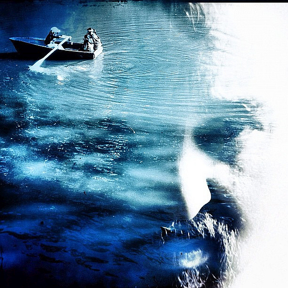 Blue monochromatic image of three people in rowboat on calm water with man's face subtly appearing in the surface of the water
